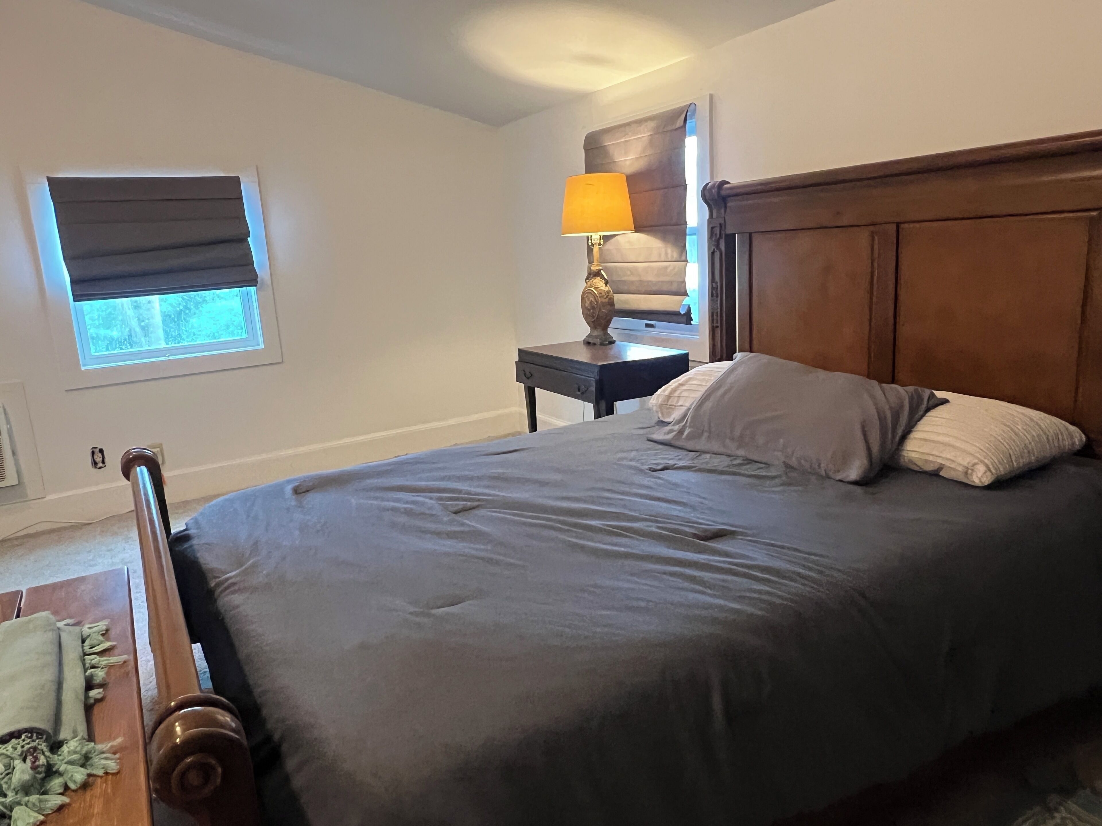Ein Schlafzimmer mit einem großen Bett, einem Nachttisch mit Lampe, einem Fenster mit Jalousien und einem Holz-Kopfteil.
