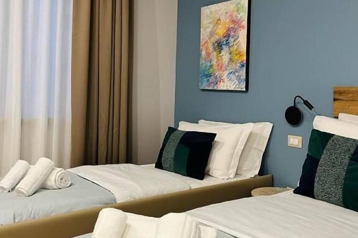 A hotel room with two beds, a blue accent wall, a colorful abstract painting, and a bedside lamp.