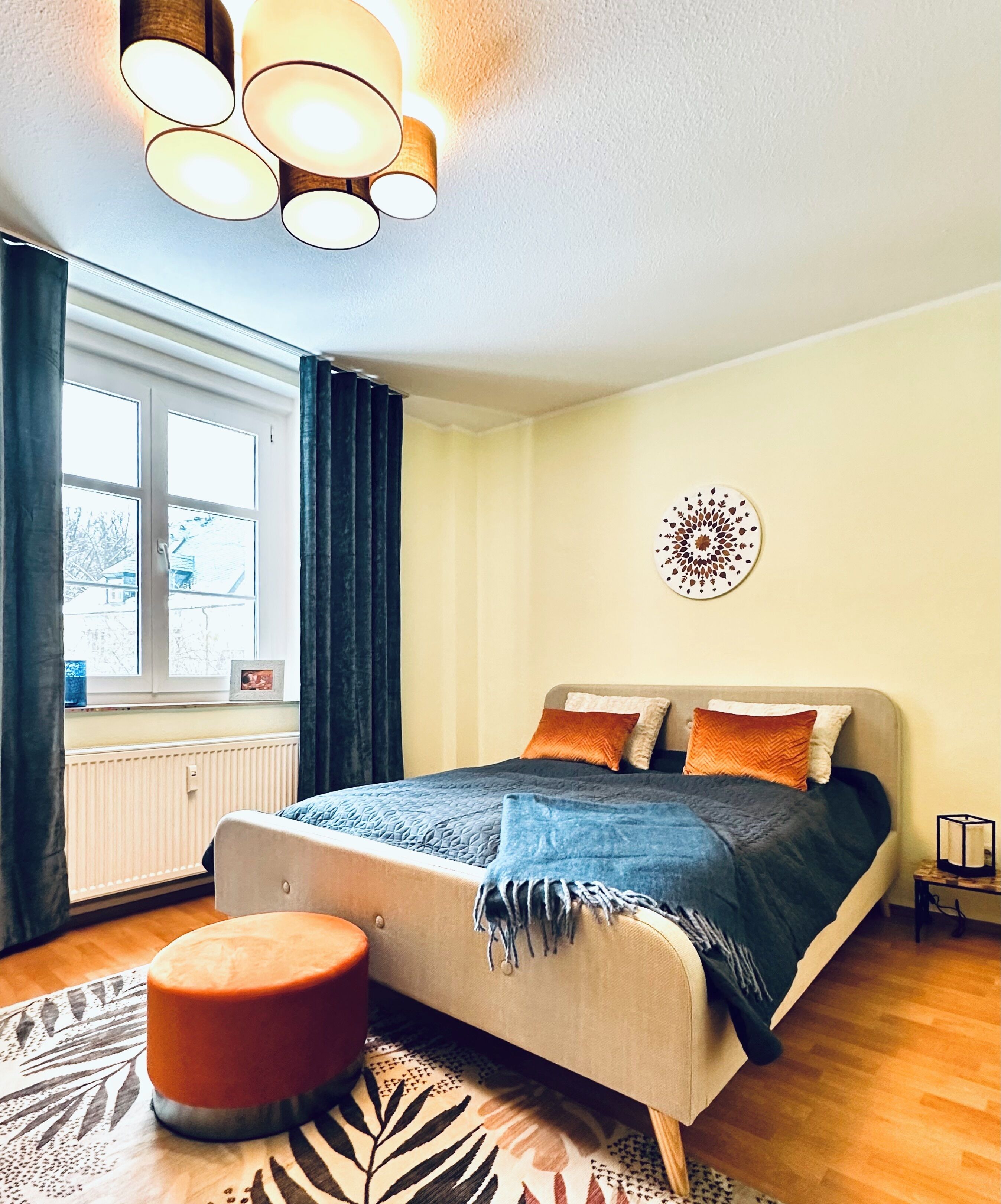 Un dormitorio con cama, ventana, radiador, una decoración en la pared y una mesita de noche con lámpara.