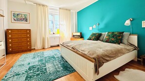 A bedroom with a bed, a dresser, a nightstand, a lamp, a window with curtains, and a painting on the wall. - Stylish apartment right by the forest, quiet and close to the center (Chemnitz)
