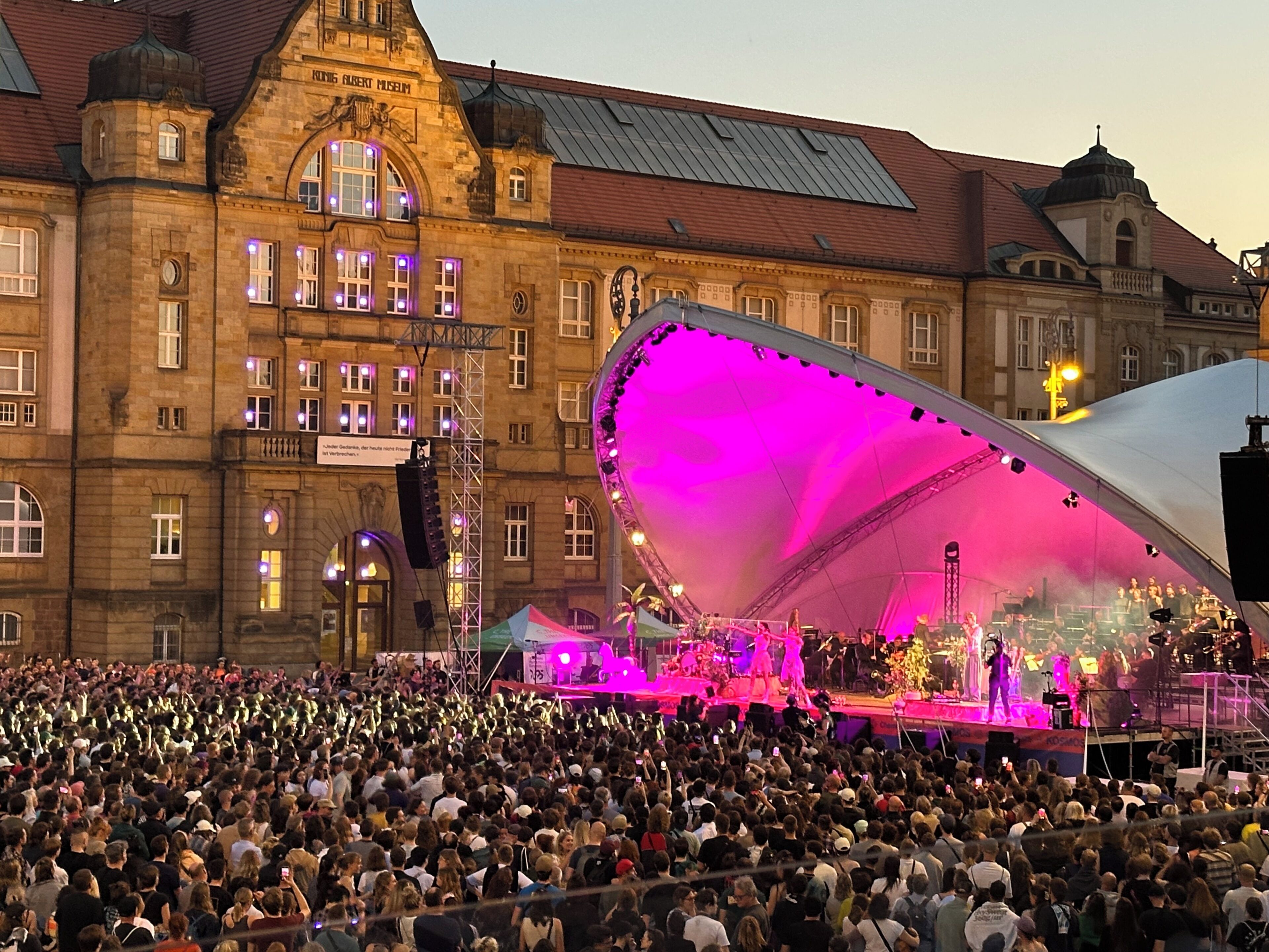 A large crowd gathered in front of a historic building with a stage set up for an event.