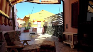 Eine sonnendurchflutete Terrasse mit einer gepolsterten Liege, einem kleinen Tisch und einem Topfpflanzen.