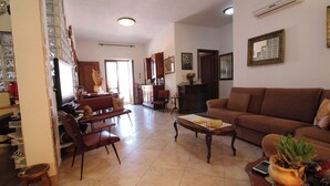 Ein Wohnzimmer mit braunem Sofa, einem Holztisch und einem Fernseher. Es gibt eine Küchenzeile mit Schränken und einem Kühlschrank.