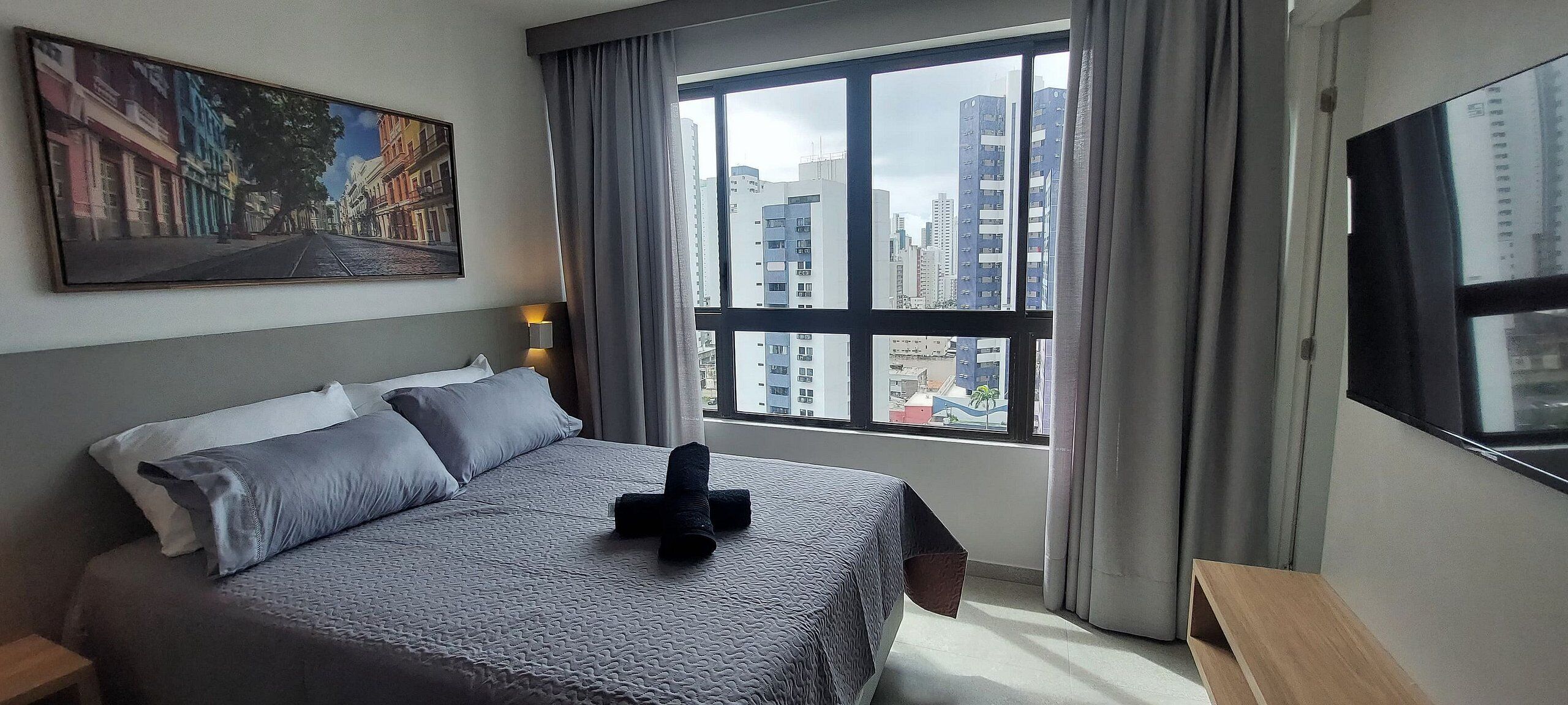 Ein Hotelzimmer mit Bett, einem Bild an der Wand, einem Fenster mit Stadtblick und einem Nachttisch.