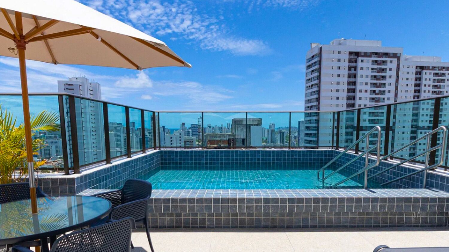 A rooftop pool with glass railings, a table with chairs, and a view of city buildings.