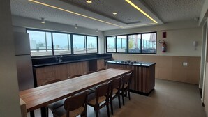 Une cuisine moderne avec une longue table et des chaises en bois, de grandes fenêtres et un extincteur fixé au mur.
