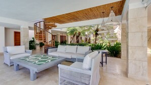 A spacious living area with a large sectional sofa, a coffee table, and a white wicker chair. A wooden staircase leads to an upper level, and there is a hanging light fixture above. - Villa Monica: Private retreat near golf w/ staff, pool & golf cart (Punta Cana)