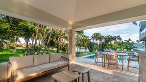 A modern outdoor living area with a sofa, dining table, and chairs, overlooking a pool and lush greenery. - Buganvilla Mansion: 2 amazing lakefront villas w/ pools, gym, games, & staff (Punta Cana)