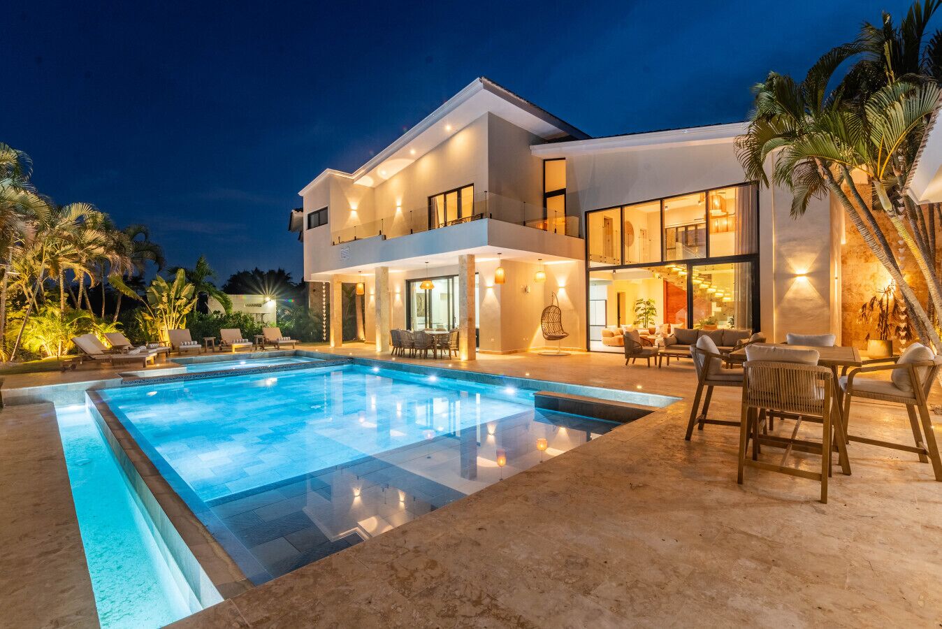Et moderne hus med to etager, swimmingpool, udendørs møbler og palmer.