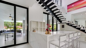 A modern kitchen with a marble countertop, a bar area with stools, a staircase, and a view of an outdoor pool area. - Las Palmas 3: Luxury retreat w/ pool, Jacuzzi, games, golf carts & full staff (Punta Cana)