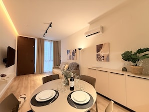 A modern dining area with a round table set for two, a sofa in the background, and a TV mounted on the wall. - VidaCalma Apto 3 (Valencia)