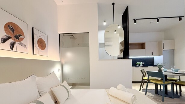 Quarto de hotel moderno com uma cama grande, área de refeições e cozinha ao fundo.