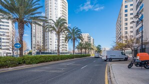 Rua urbana com edifícios residenciais altos, palmeiras e uma carrinha branca estacionada ao lado.