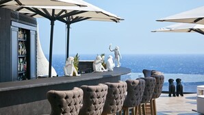A bar area with a view of the ocean, featuring bar stools and umbrellas. - Royal Myconian - Leading Hotels Of The World (Mykonos)