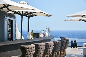 A bar area with a view of the ocean, featuring bar stools and umbrellas. - Royal Myconian - Leading Hotels Of The World (Mykonos)