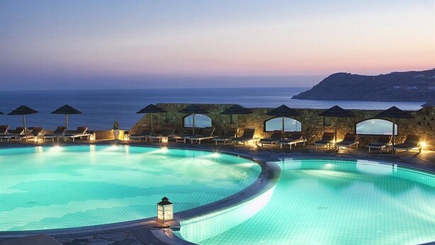 A large, curved swimming pool with lounge chairs and umbrellas, surrounded by stone walls, overlooking the sea. - Royal Myconian - Leading Hotels Of The World (Mykonos)