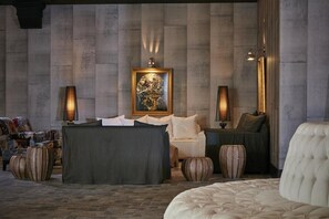 A modern lounge area with a sofa, armchairs, and a coffee table. There is a framed artwork on the wall and a lamp on each side of the sofa. - Royal Myconian - Leading Hotels Of The World (Mykonos)