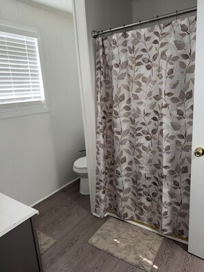 A bathroom with a toilet, a shower curtain with a leaf pattern, a window with blinds, and a wooden floor.
