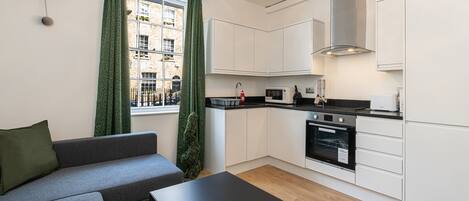 Una cocina moderna con armarios blancos, encimera negra y horno empotrado. En la sala de estar hay un sofá gris y una mesa negra.
