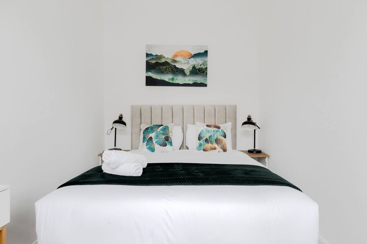 Een net opgemaakt bed met witte lakens, een donkergroene deken en twee sierkussens met een natuuristisch design.