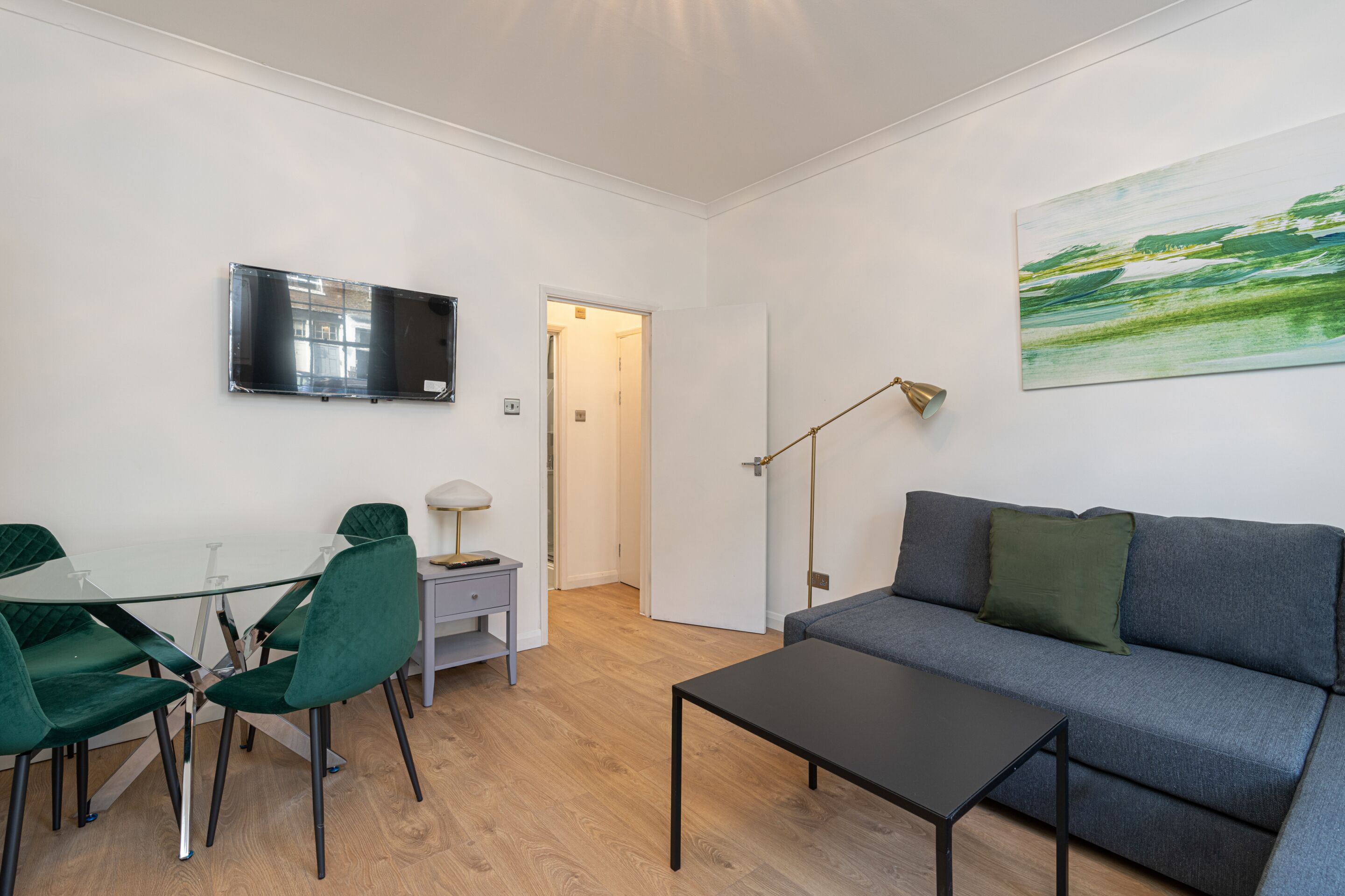 Una sala moderna con una mesa de comedor de cristal, sillas verdes, un sofá gris, una mesa de centro negra, un televisor montado en la pared y un cuadro.