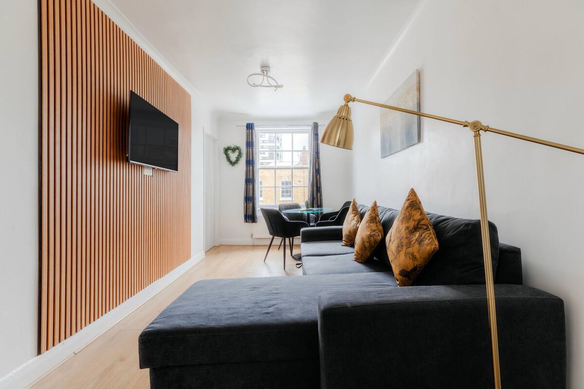 Een moderne woonkamer met een modulaire bank, een flatscreen televisie bevestigd aan een houten wandpaneel, een eethoek met tafel en stoelen, en een groot raam met gordijnen.