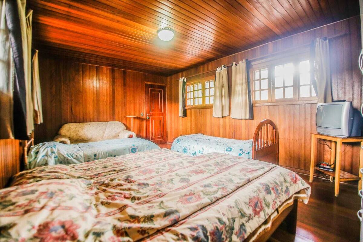 Ein Zimmer mit Holzwänden und -decke, zwei Betten mit Blumenoptik, ein Holzstuhl, ein Fernseher auf einem Holzboden und ein Fenster mit Vorhängen.