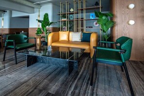 A modern lobby with a leather sofa, green armchairs, a marble coffee table, and wooden shelving with plants.