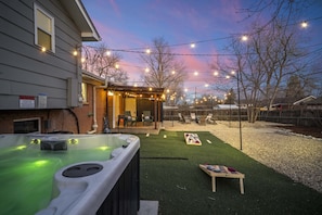 Een achtertuin met een hottub, kunstgras, een houten tafel en lichtjes.