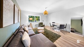 Ein modernes Wohnzimmer mit einer Couch, einem Esstisch und Stühlen. Ein Fenster bietet Blick auf Grünflächen.