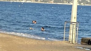 En strand med tre personer som badar i vattnet, ett livräddartorn och en sandstrand.