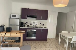 Une cuisine compacte avec des meubles de couleur violette, un four à micro-ondes, un grille-pain et un évier. On y trouve une table à manger avec des chaises, ainsi qu’une petite table ronde sur laquelle se trouve un ordinateur portable.