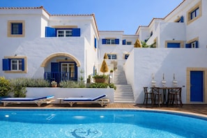 Zona de piscina amb una piscina blava cristal·lina, edificis blancs amb detalls blaus i una escala que condueix a un nivell superior.