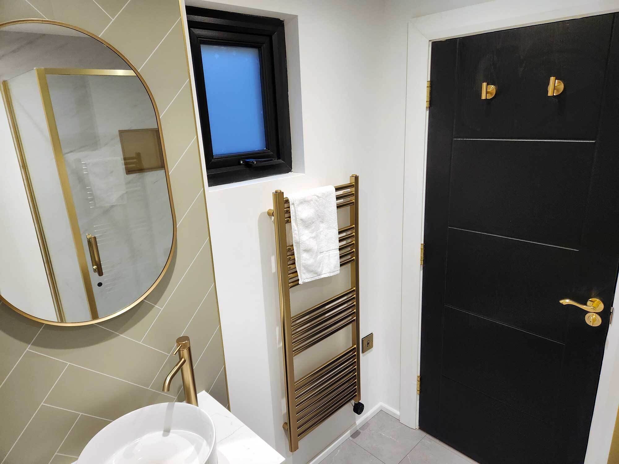 A bathroom with a white sink, a gold-framed mirror, a towel rack with a white towel, a black door with gold handles, and a small window with a blue view.