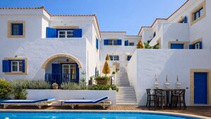 A poolside area with a clear blue pool, white buildings with blue accents, and a staircase leading to an upper level.