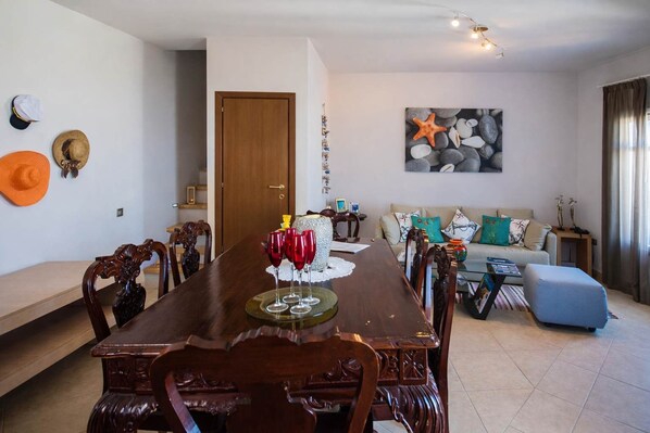 A dining area with a wooden table, chairs, and a decorative centerpiece. There is a sofa and a painting on the wall in the background.