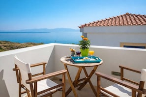 A balcony with a table set for breakfast, a potted plant, and a view of the sea and mountains.