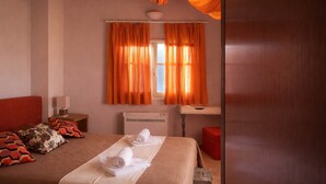 A bedroom with a bed, two rolled towels, a nightstand with a lamp, a window with orange curtains, and a wooden ceiling. - Castri Village Kythira - Phoenix Sea View Retreat (Agia Pelagia)