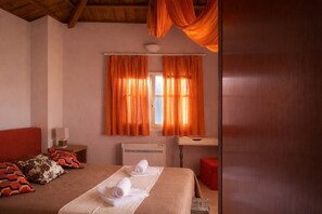 A bedroom with a bed, two rolled towels, a nightstand with a lamp, a window with orange curtains, and a wooden ceiling.