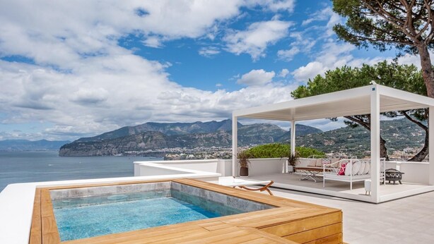 Un espace piscine extérieur moderne, doté d’une terrasse en bois, d’un coin salon couvert offrant une vue sur la mer et les montagnes, et d’un ciel dégagé.