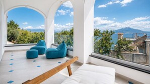 Une pièce moderne avec un canapé blanc, des fauteuils en forme de poire bleus et un banc en bois. De grandes fenêtres offrent une vue sur un paysage montagneux et un plan d’eau.