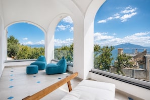 Ein modernes Zimmer mit einem weißen Sofa, blauen Sitzsäcken und einer Holzbank. Große Fenster bieten Blick auf eine Berglandschaft und ein Gewässer.