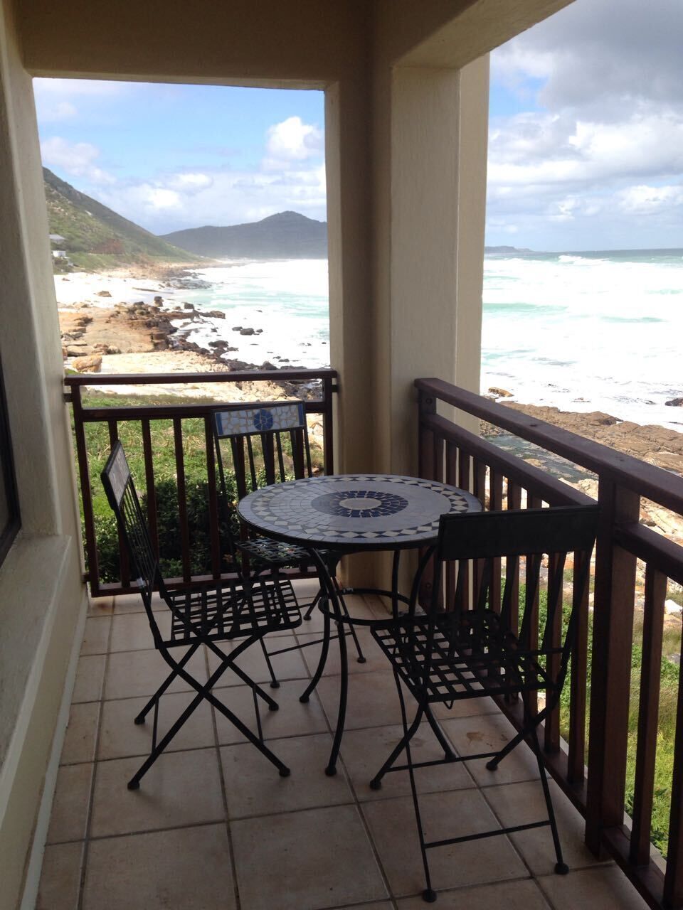 Ein Balkon mit rundem Tisch und zwei Stühlen, der Blick auf einen Strand mit Wellen und einen entfernten Berg freigibt.