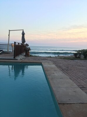 A swimming pool adjacent to a beach with waves, a wooden deck, and a metal fence. - Fabulous four bedroomed beach house in a spectacular coastal setting. (Cape Town)