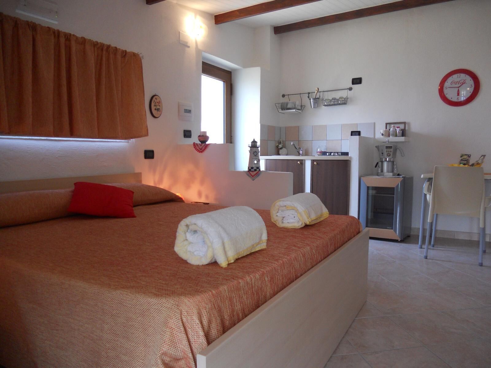 Ein Schlafzimmer mit einem Bett, einem roten Kissen, zwei aufgerollten Handtüchern, einem Fenster, einer Küchenzeile mit Spüle, einer Kaffeemaschine und einer Uhr an der Wand.