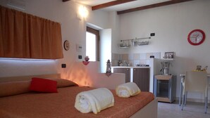 A bedroom with a bed, a red pillow, two rolled towels, a window, a kitchen area with a sink, a coffee machine, and a clock on the wall. - Relaxation, nature and history in the historic village a 5 km from the sea. (Ogliastro Cilento)