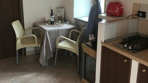 A compact kitchen with a gas stove, a small dining table with chairs, a window with curtains, and a wall-mounted light fixture. - Relaxation, nature and history in the historic village a 5 km from the sea. (Ogliastro Cilento)