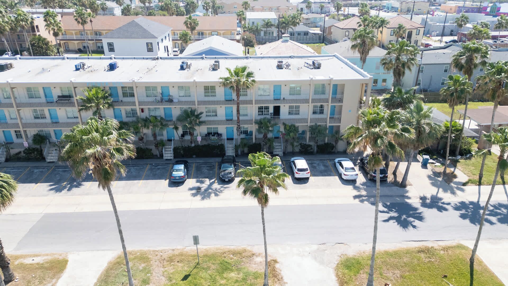 A multi-story hotel with a flat roof, several parking spaces, and palm trees in front.