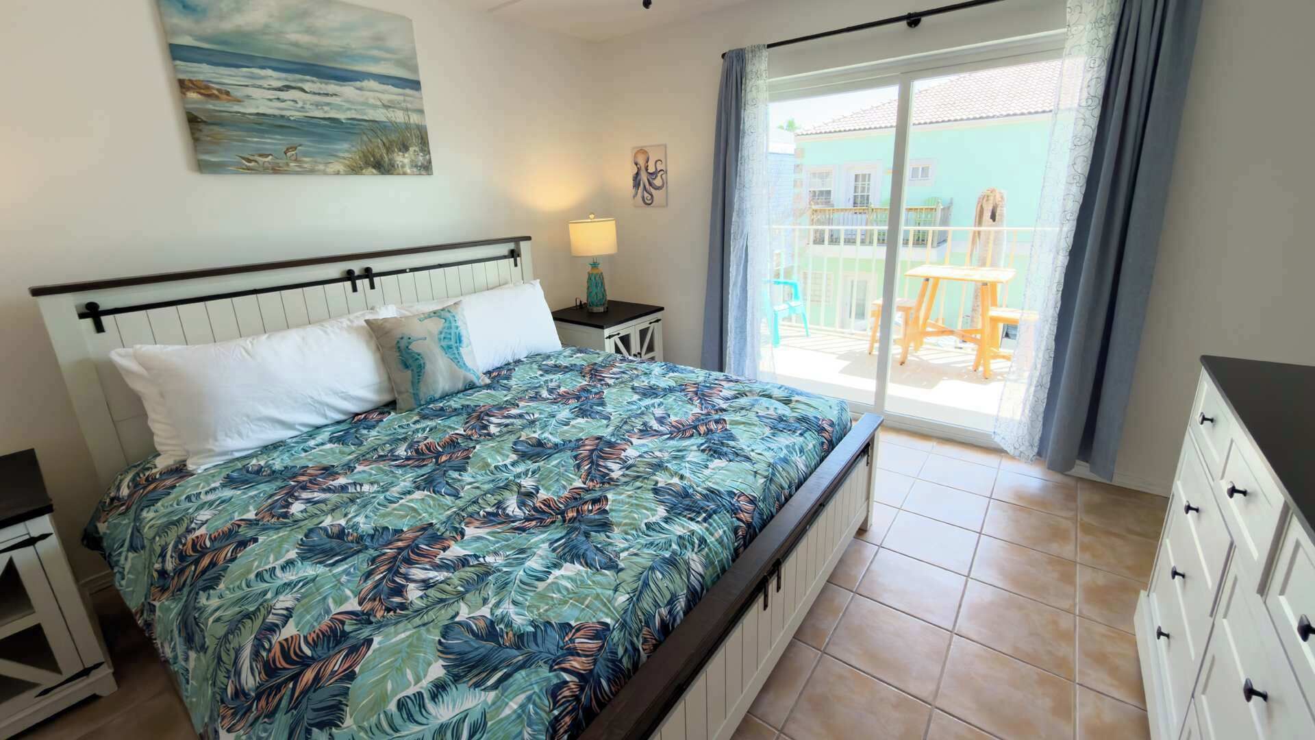 Una camera da letto con un letto, un comodino, una cassettiera, un quadro e vista sul balcone esterno.