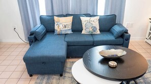 Sala de estar com um sofá modular azul, duas almofadas decorativas, uma mesa de centro redonda e uma mesinha lateral.